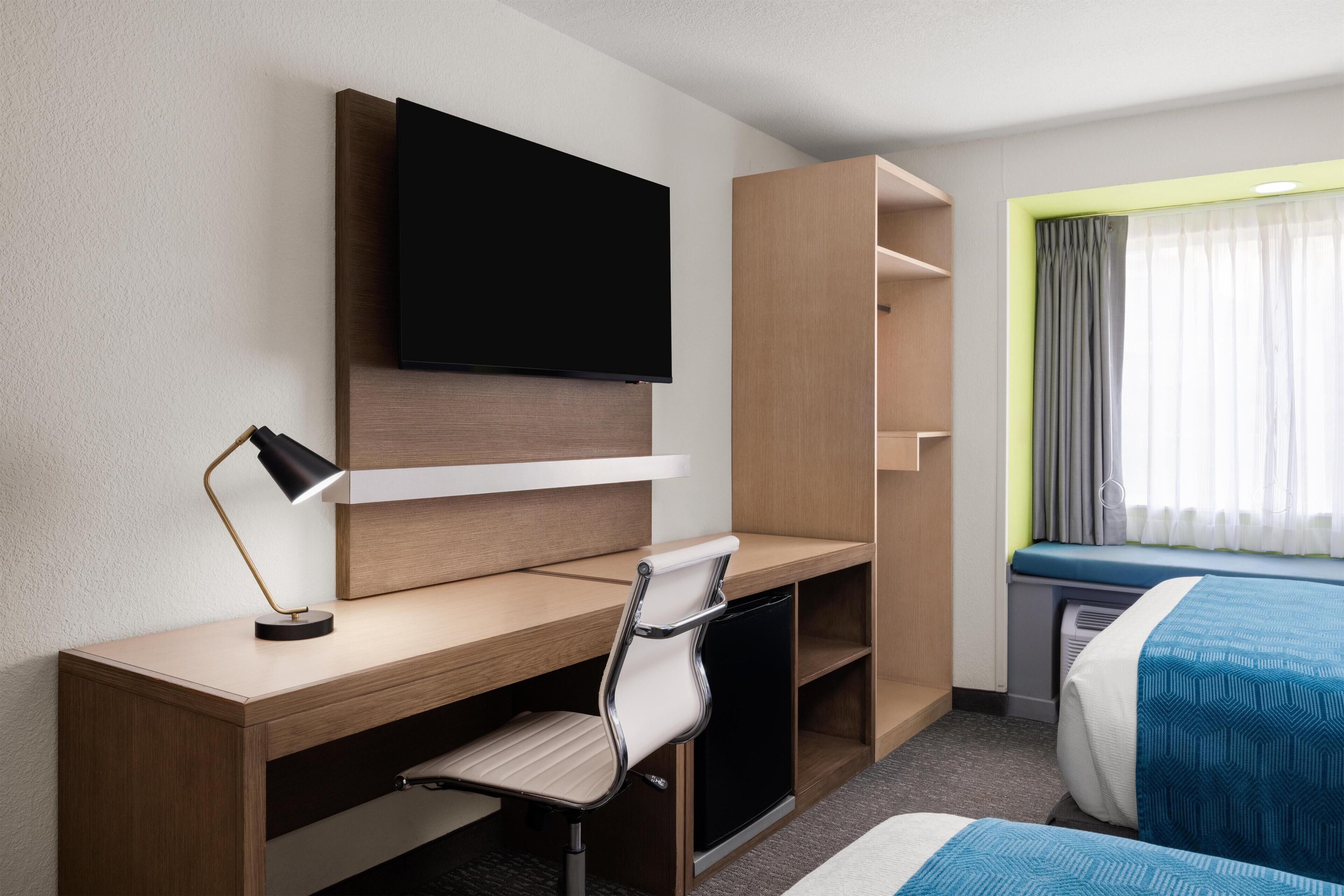 A hotel room with a wooden desk, a chair, a television mounted on the wall, a bed with blue bedding, and a window with curtains.