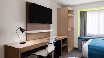 Habitación de hotel con escritorio de madera, una silla, un televisor montado en la pared, cama con ropa de cama azul y una ventana con cortinas.