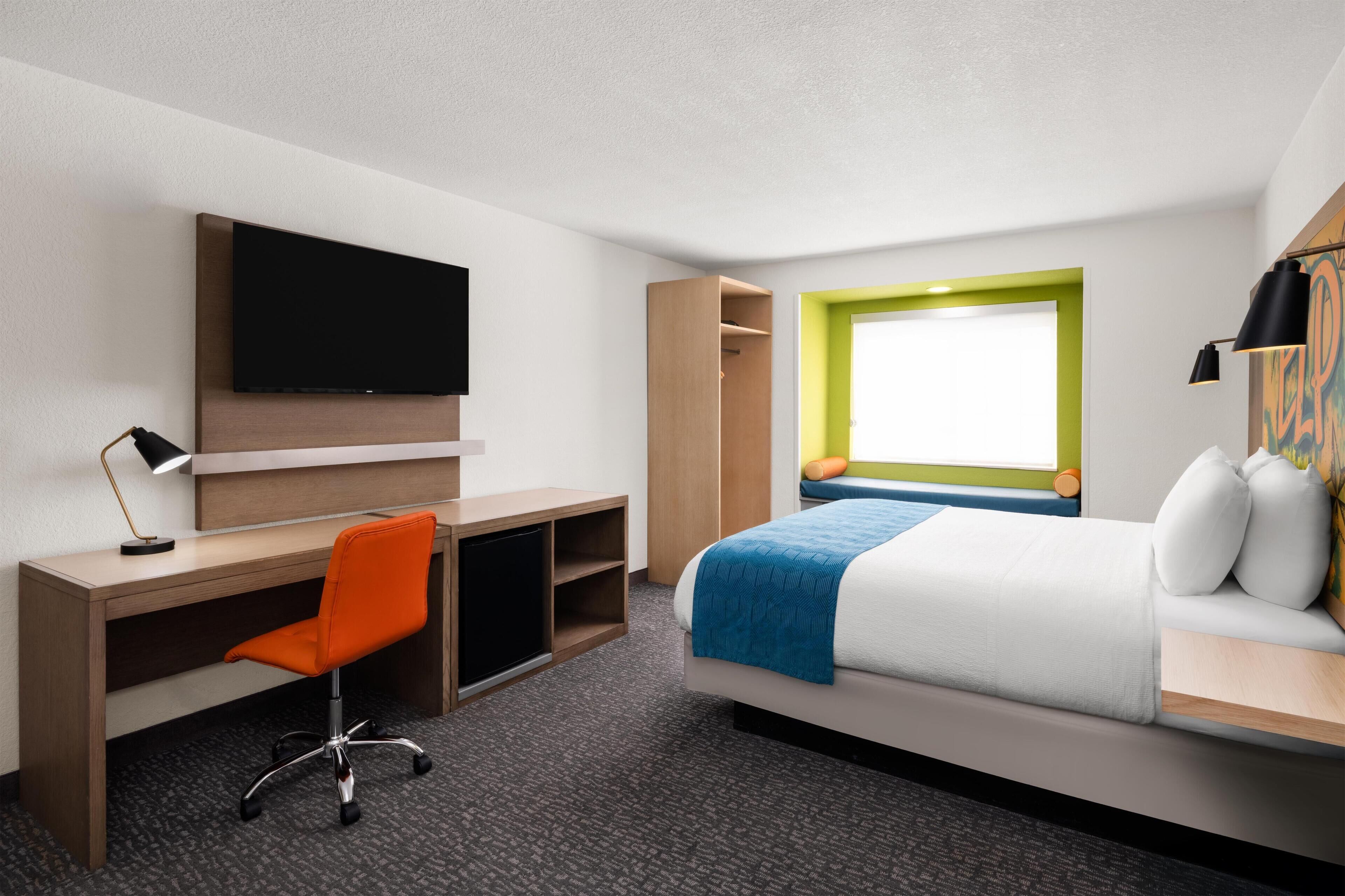 A hotel room with a bed, a desk, a chair, a TV, a wardrobe, and a window with a green frame.