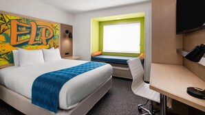 A hotel room with a bed, a desk, a TV, a window with a view, and a colorful wall decoration.