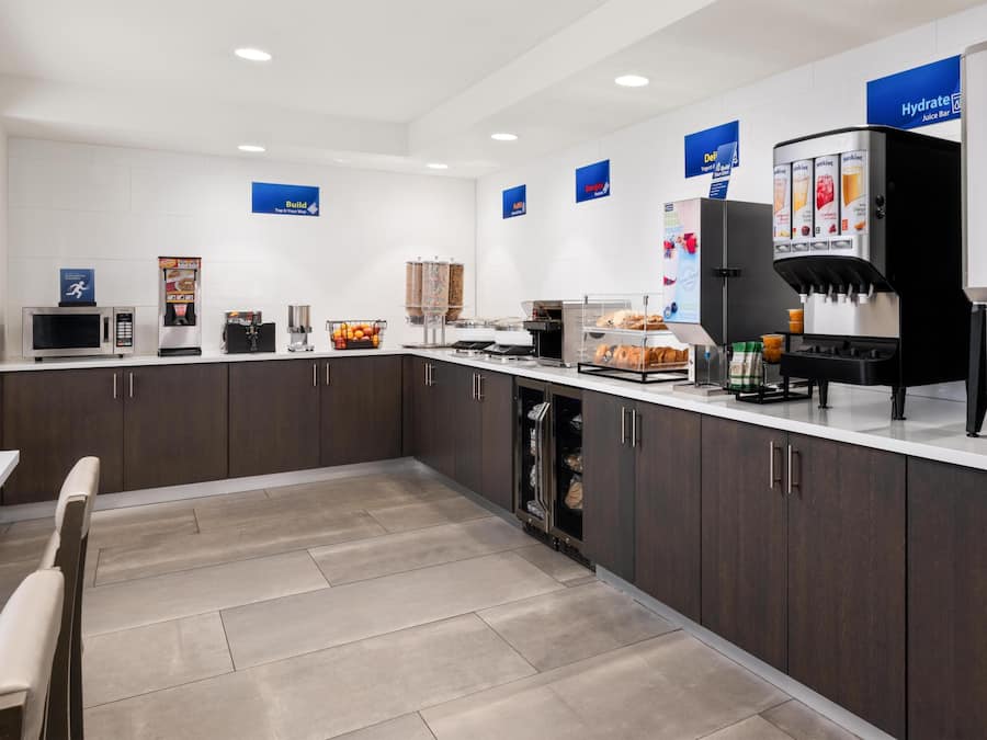 A modern kitchen with a microwave, coffee machine, and juice dispenser. There are cabinets, a refrigerator, and a counter with various items.