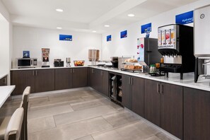 Una cocina moderna con microondas, cafetera y dispensador de jugo. Hay armarios, un refrigerador y una encimera con varios artículos.