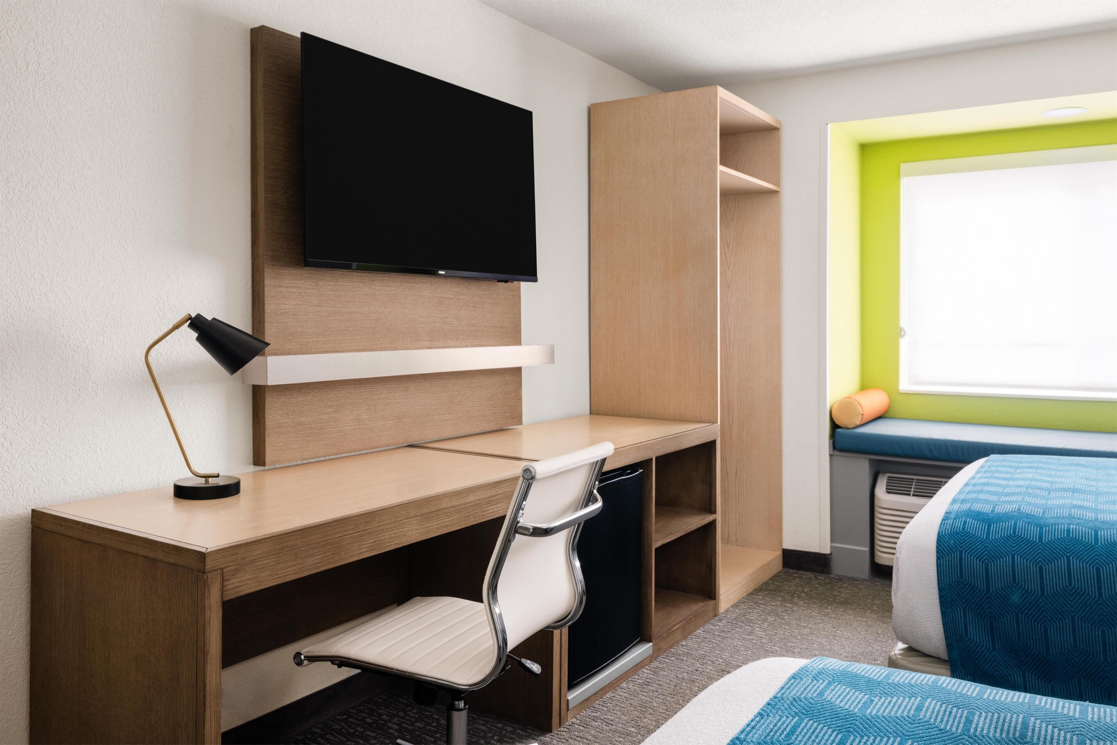 A hotel room with a wooden desk, a chair, a television mounted on the wall, a bed with a blue and white patterned bedspread, and a window with a green frame.