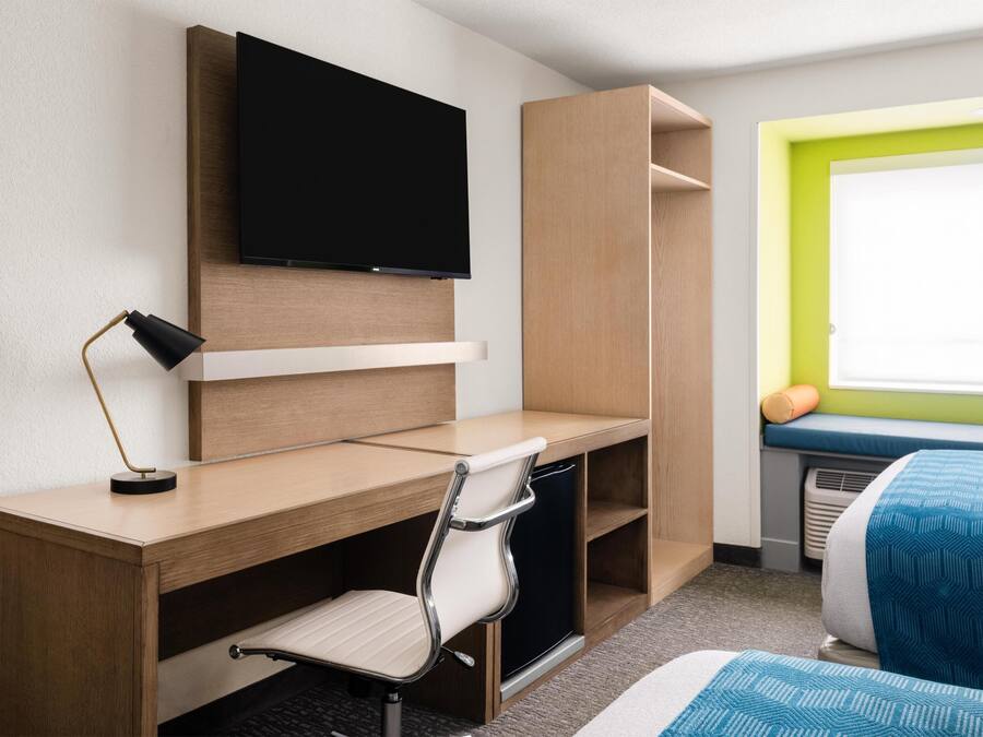 A hotel room with a wooden desk, a chair, a television mounted on the wall, a bed with a blue and white patterned bedspread, and a window with a green frame.