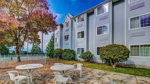 A two-story building with a patio area featuring round tables and chairs, surrounded by trees and shrubs. - Motel 6 Sutherlin, OR (Sutherlin)
