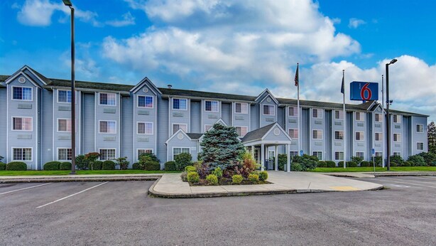 A motel with a central entrance, a parking lot, and a large evergreen tree in the foreground. - Motel 6 Sutherlin, OR (Sutherlin)