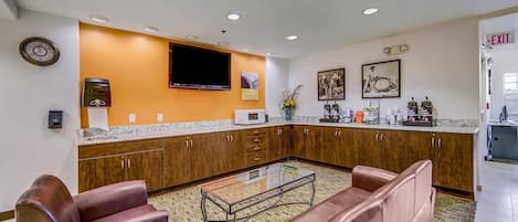 Una sala de estar moderna con sofás de cuero, una mesa de centro de vidrio, armarios de madera, un televisor de pantalla plana y una zona de cocina con microondas y cafetera.