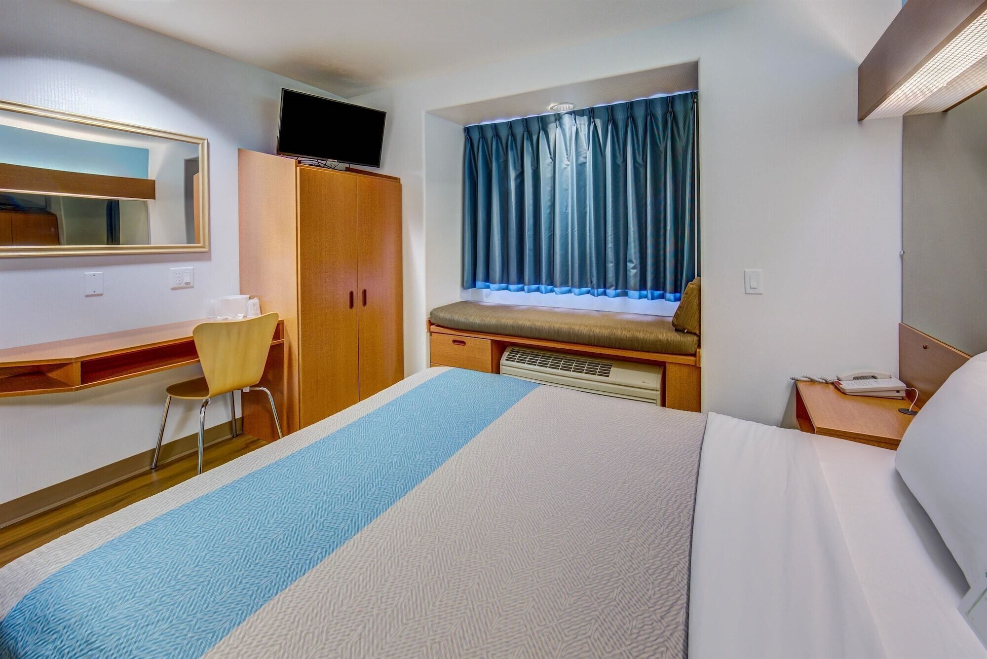 A hotel room with a bed, a desk, a chair, a wardrobe, a window with curtains, and a television on the wall.