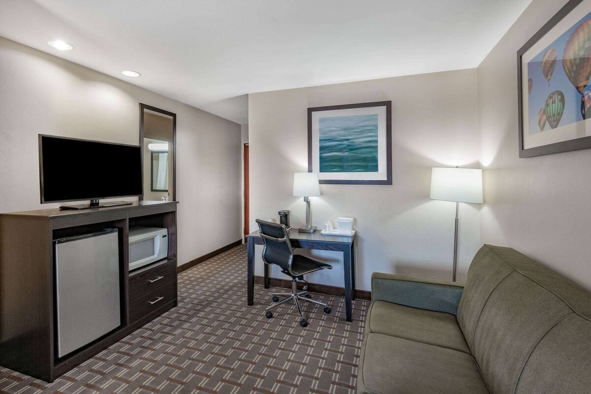 A hotel room with a flat-screen TV, a desk with a chair, a microwave, a refrigerator, a sofa, and two wall-mounted lamps.