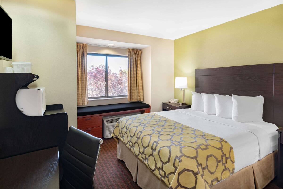 A hotel room with a bed, a desk, a chair, a window with curtains, and a lamp.