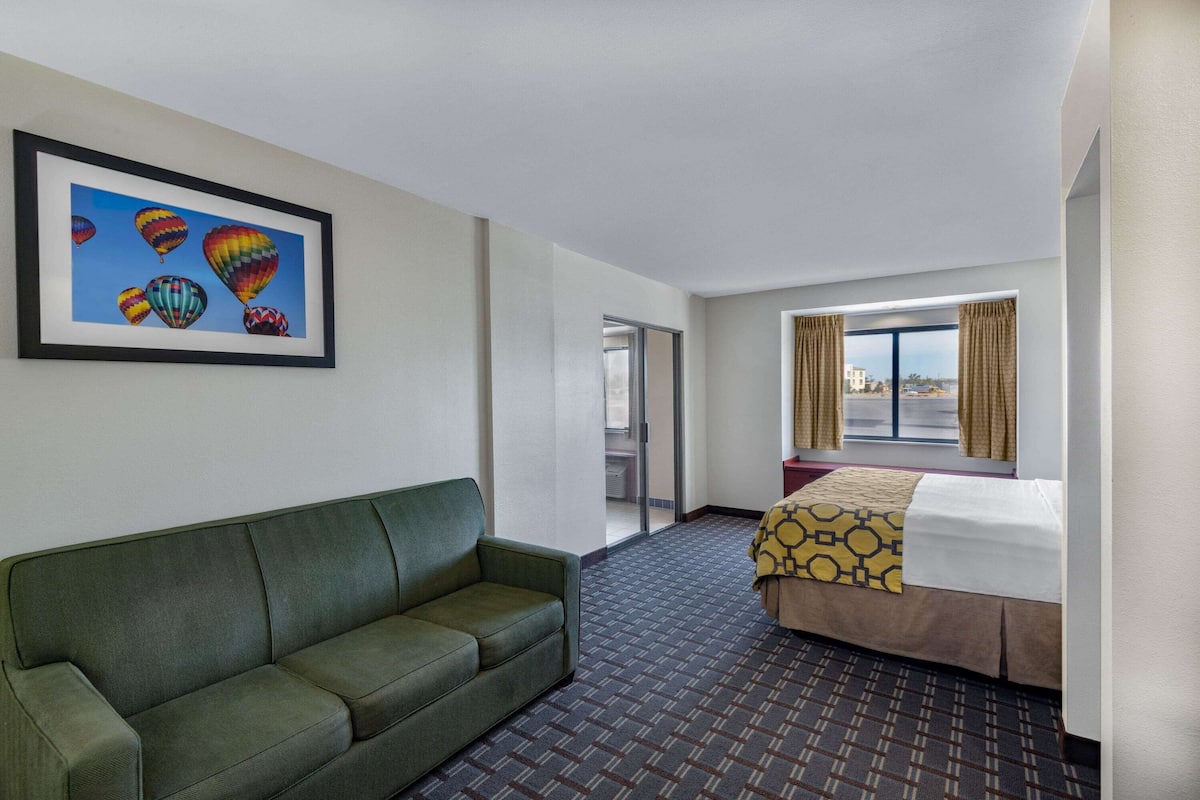 A hotel room with a green sofa, a bed with a patterned bedspread, a large window with curtains, and a framed picture of hot air balloons on the wall.