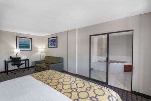 A hotel room with a large bed, a desk with a chair, a sofa, and a sliding glass door leading to a bathroom with a bathtub. - Baymont Inn by Wyndham Farmington (Farmington)