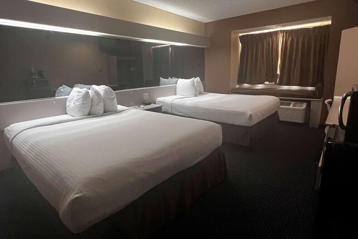 A hotel room with two beds, a large mirror, a window with curtains, and a desk with a lamp.