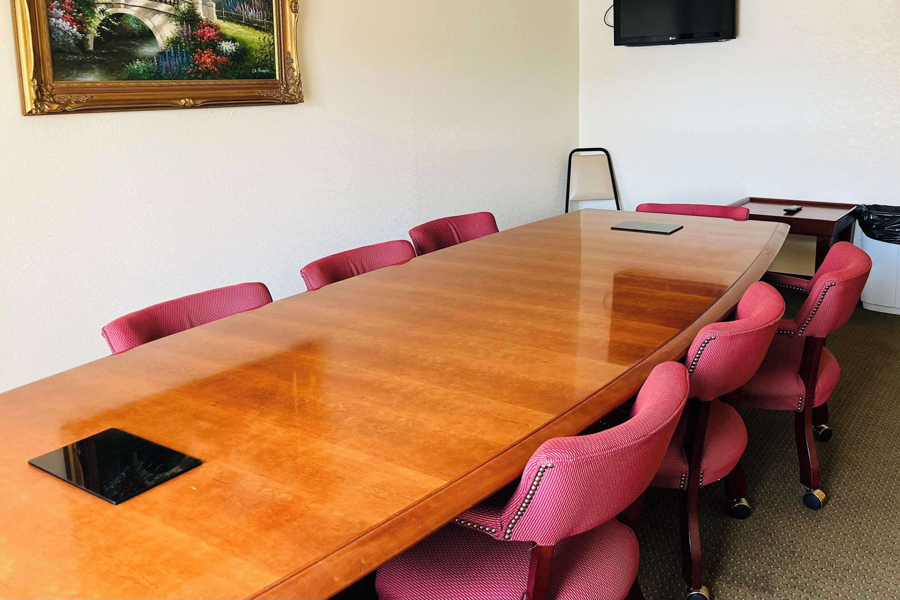 A conference room with a long wooden table, red upholstered chairs, a framed painting on the wall, and a television mounted on the wall.