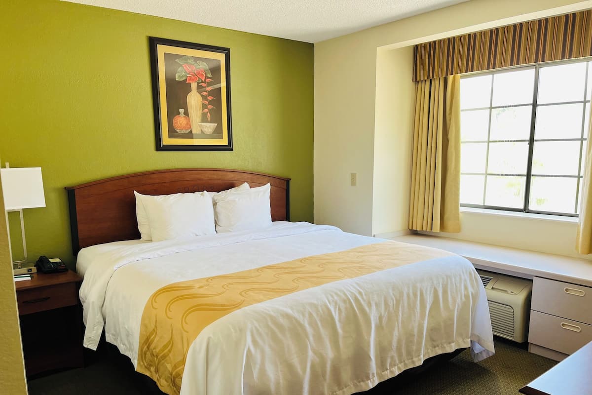 A hotel room with a bed, a nightstand, a desk, a window with curtains, and a painting on the wall.
