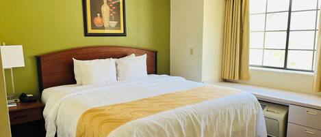Una habitación de hotel con una cama, una mesita de noche, un escritorio, una ventana con cortinas y un cuadro en la pared.