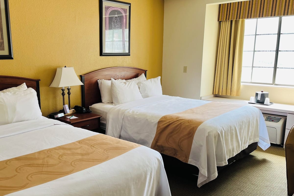 A hotel room with two beds, a bedside table with a lamp, a window with curtains, and a wall-mounted artwork.