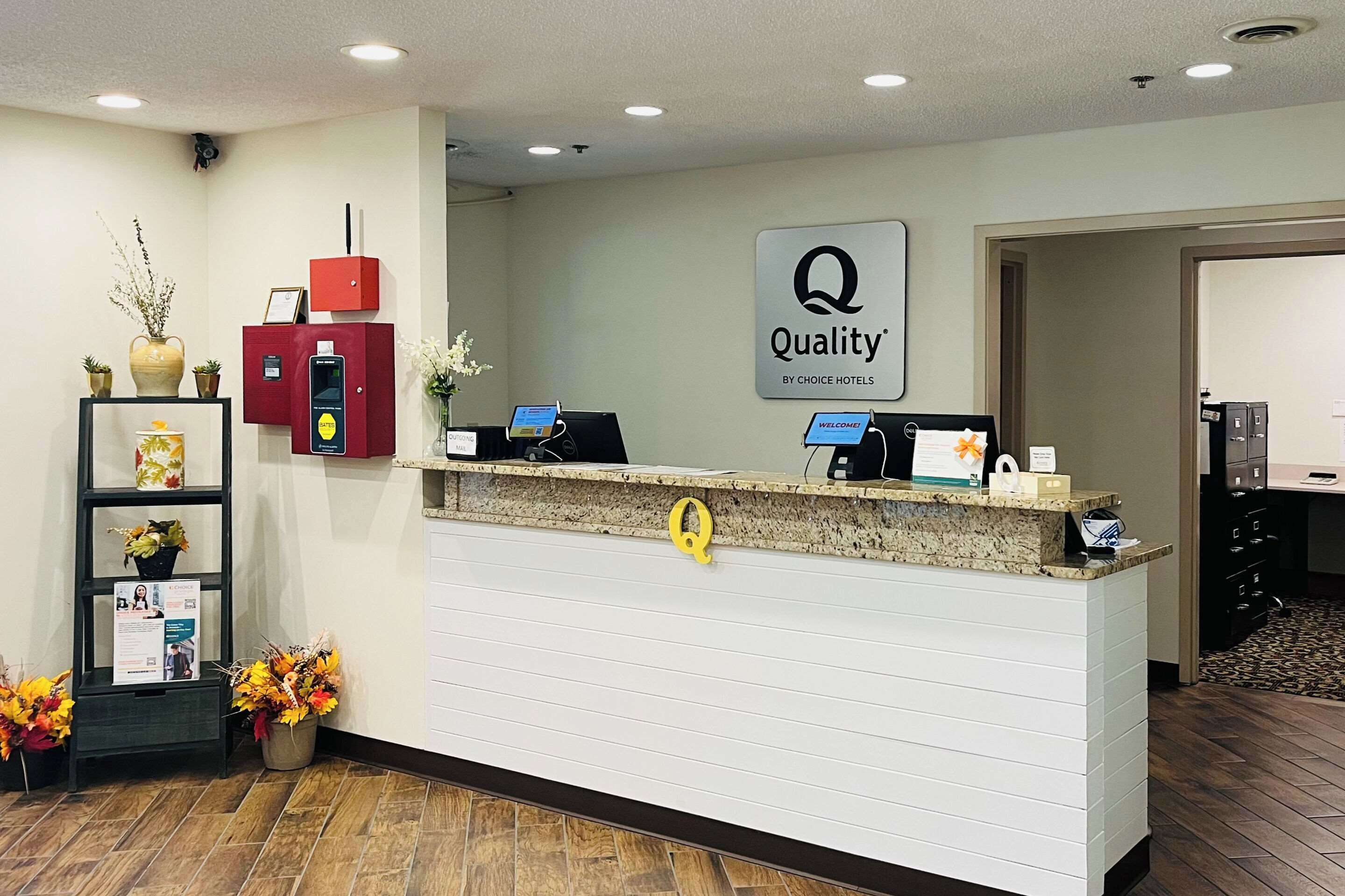 A hotel reception desk with a 'Quality by Choice Hotels' sign, a red information kiosk, and a plant display.