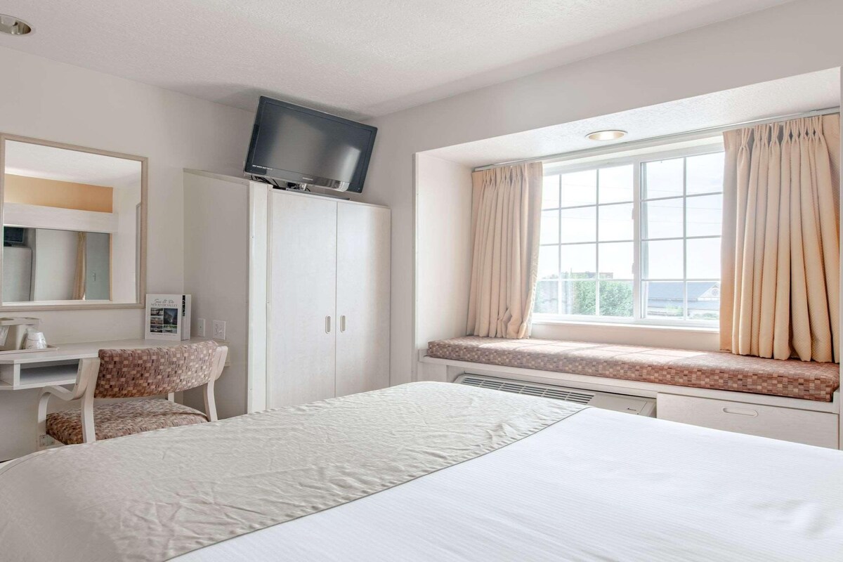 Una habitación de hotel con cama, escritorio con silla, televisor sobre un armario, ventana con cortinas y vista al exterior.