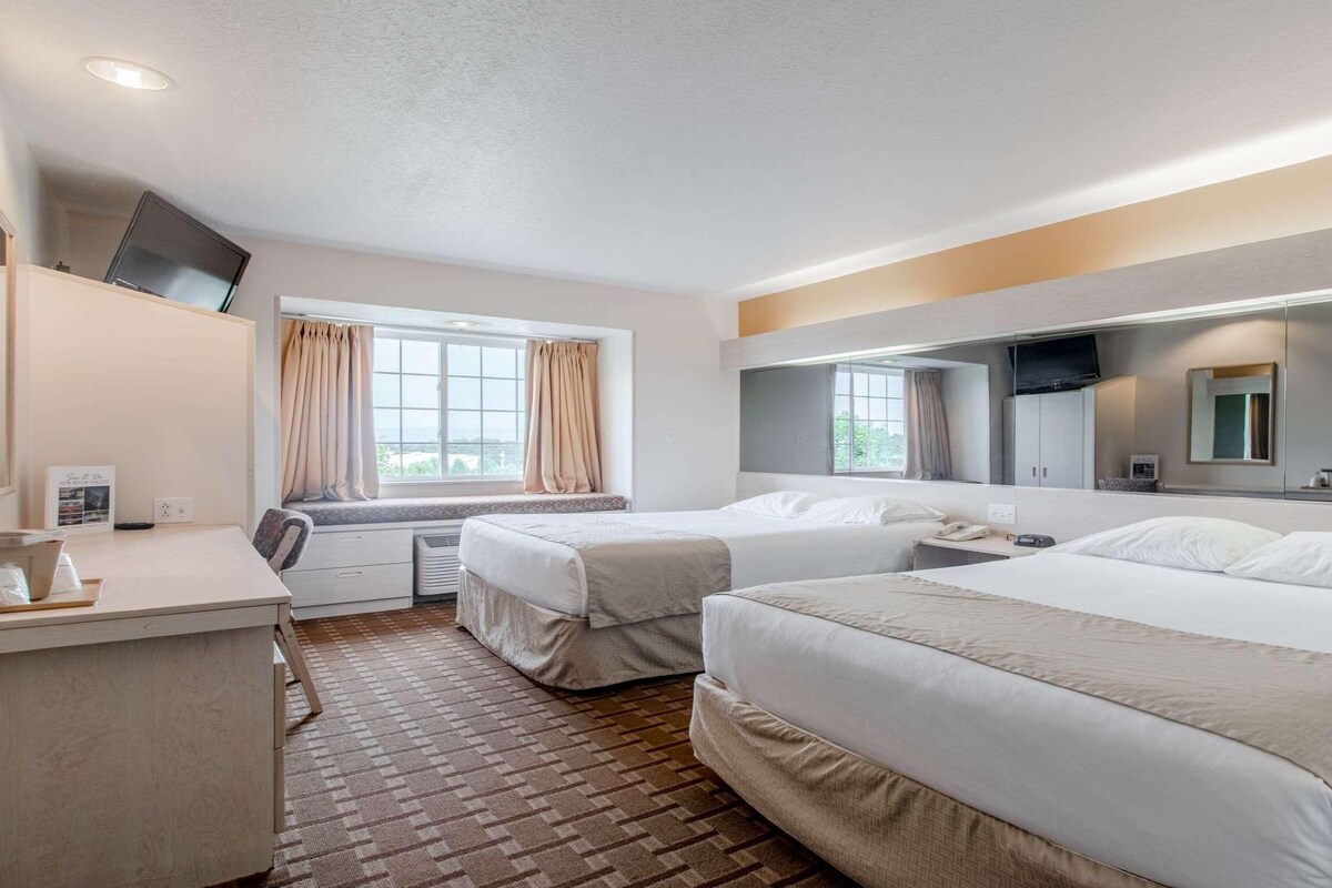 Habitación de hotel con dos camas, un escritorio, una ventana con cortinas y un televisor de pantalla plana montado en la pared.