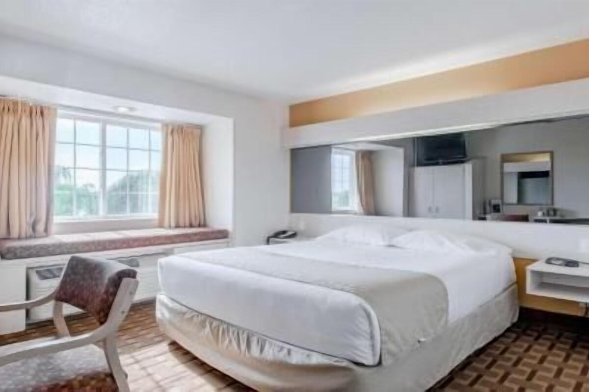 Una habitación de hotel con una cama grande, un escritorio, una silla, un ventanal con cortinas y un espejo.