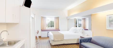 A hotel room with a bed, a sofa, a desk with a lamp, a window with curtains, and a TV mounted on the wall.
