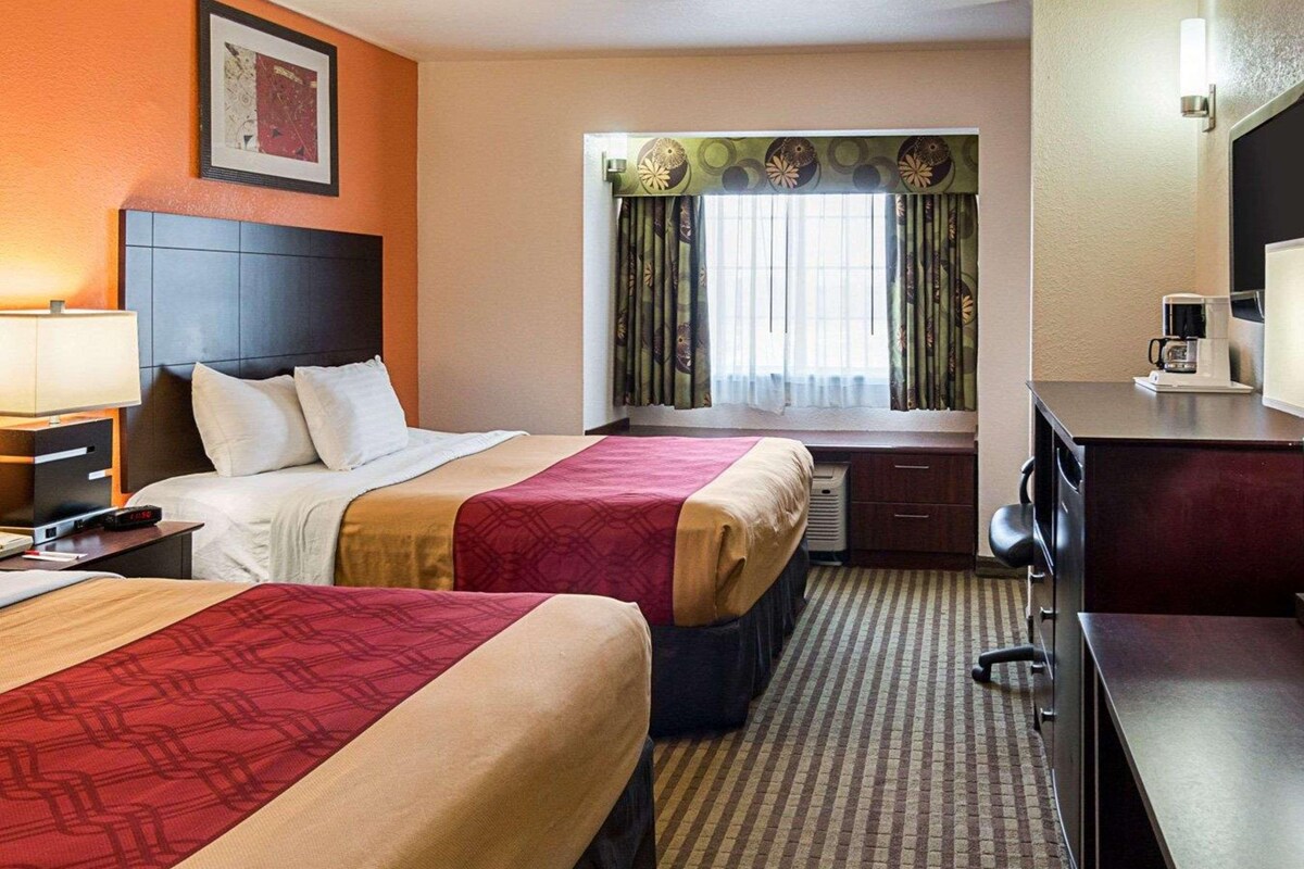 A hotel room with two beds, a desk, a chair, a lamp, a coffee maker, and a window with curtains.