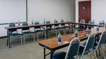 Sala de conferência com uma mesa comprida, cadeiras, garrafas de água, tela de projetor e uma porta com uma placa.
