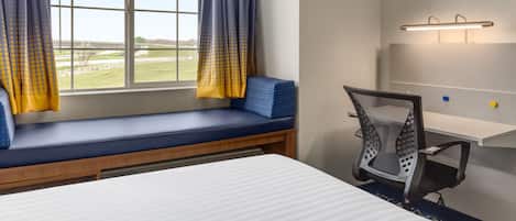 Quarto de hotel com uma janela grande, um banco com almofadas, uma mesa com cadeira e uma estante na parede.