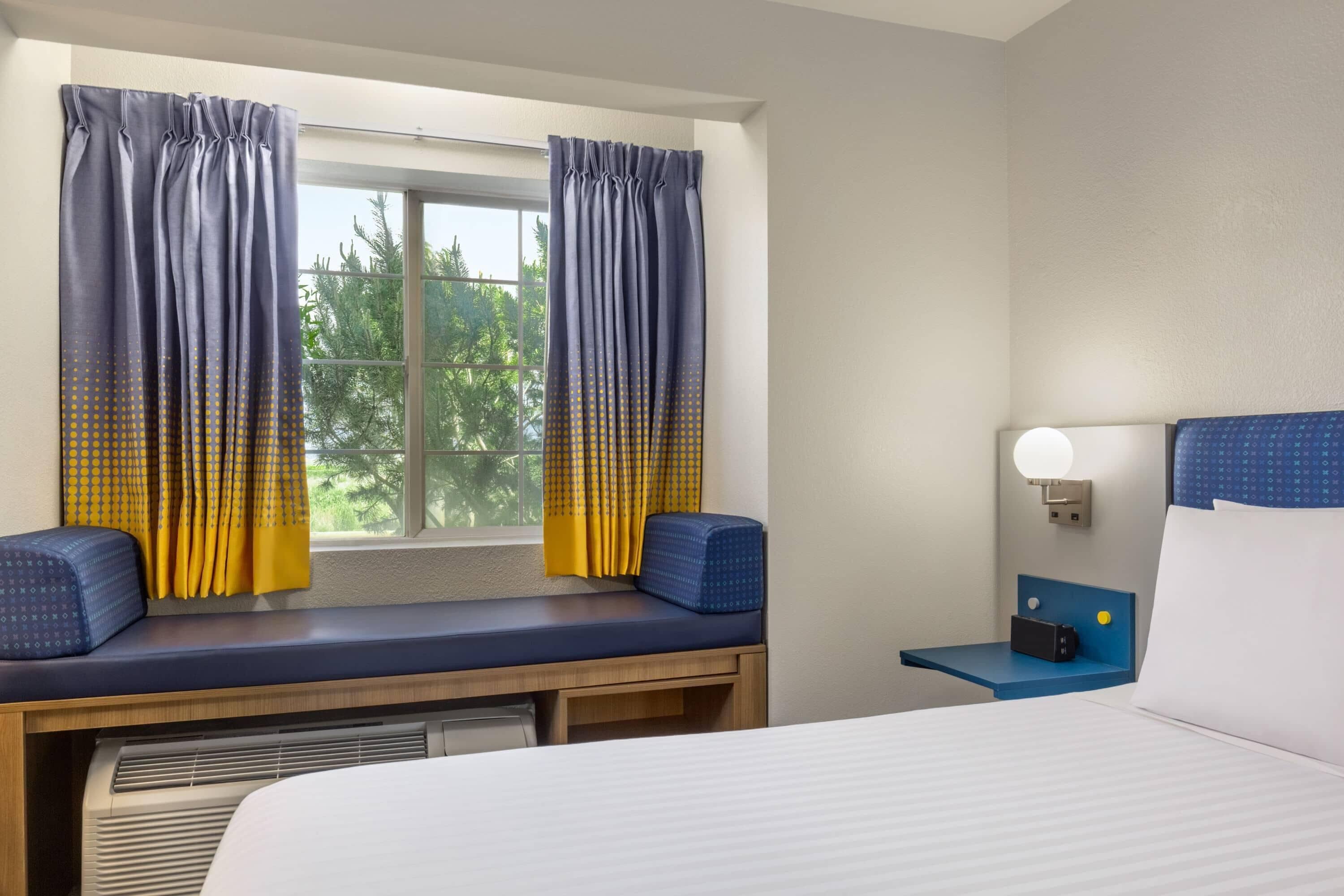A hotel room with a bed, a bedside table with a lamp, a window with curtains, and a bench with cushions.