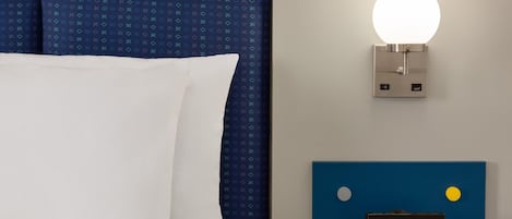 A bed with a white pillow and a blue headboard, a wall-mounted light fixture, and a small table with a black object on it.