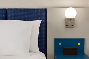 A bed with a white pillow and a blue headboard, a wall-mounted light fixture, and a small table with a black object on it.