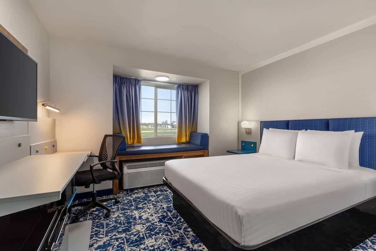 Habitación de hotel con cama, escritorio, silla, ventana con cortinas y televisión en la pared.