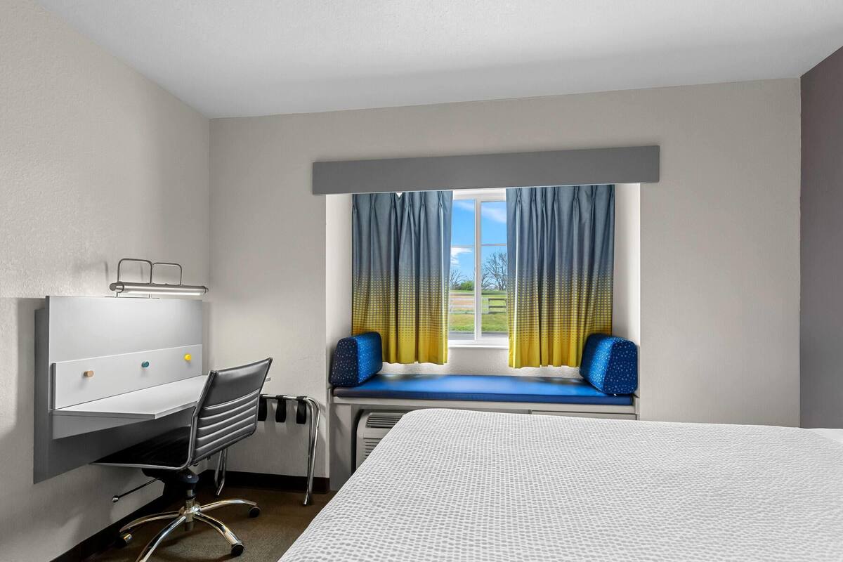 Ein Hotelzimmer mit Bett, Schreibtisch mit Stuhl, einem Fenster mit Vorhängen und Blick ins Freie.