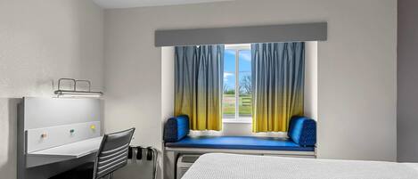 A hotel room with a bed, a desk with a chair, a window with curtains, and a view of the outdoors.