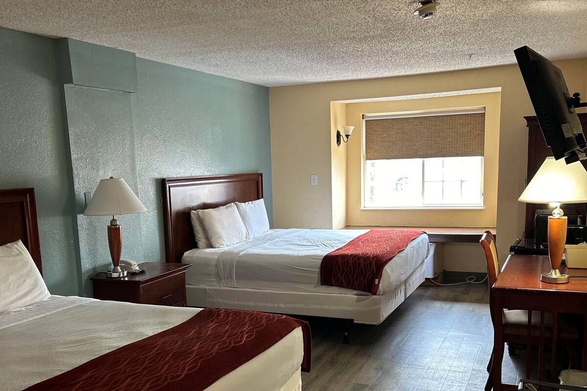 A hotel room with two beds, a desk with a lamp, a window with blinds, and a ceiling light.