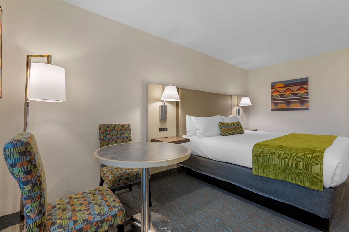 A hotel room with a bed, a round table, two patterned chairs, a nightstand, and two wall-mounted lamps.