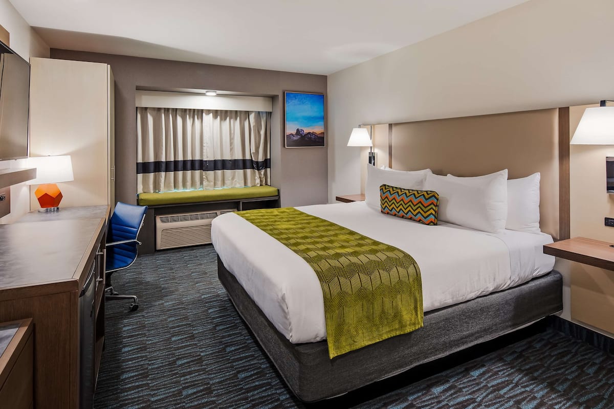 A hotel room with a bed, bedside tables, a desk, a chair, a window with curtains, and a painting on the wall.
