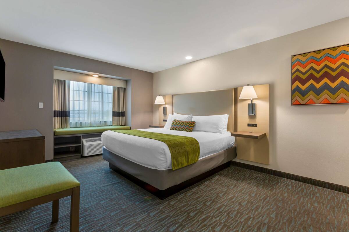 A hotel room with a bed, bedside lamps, a window with curtains, a bench, and a colorful wall art piece.
