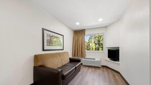 A small, furnished room with a brown leather sofa, a mounted television, and a window with curtains. - MainStay Suites Raleigh North (Raleigh)