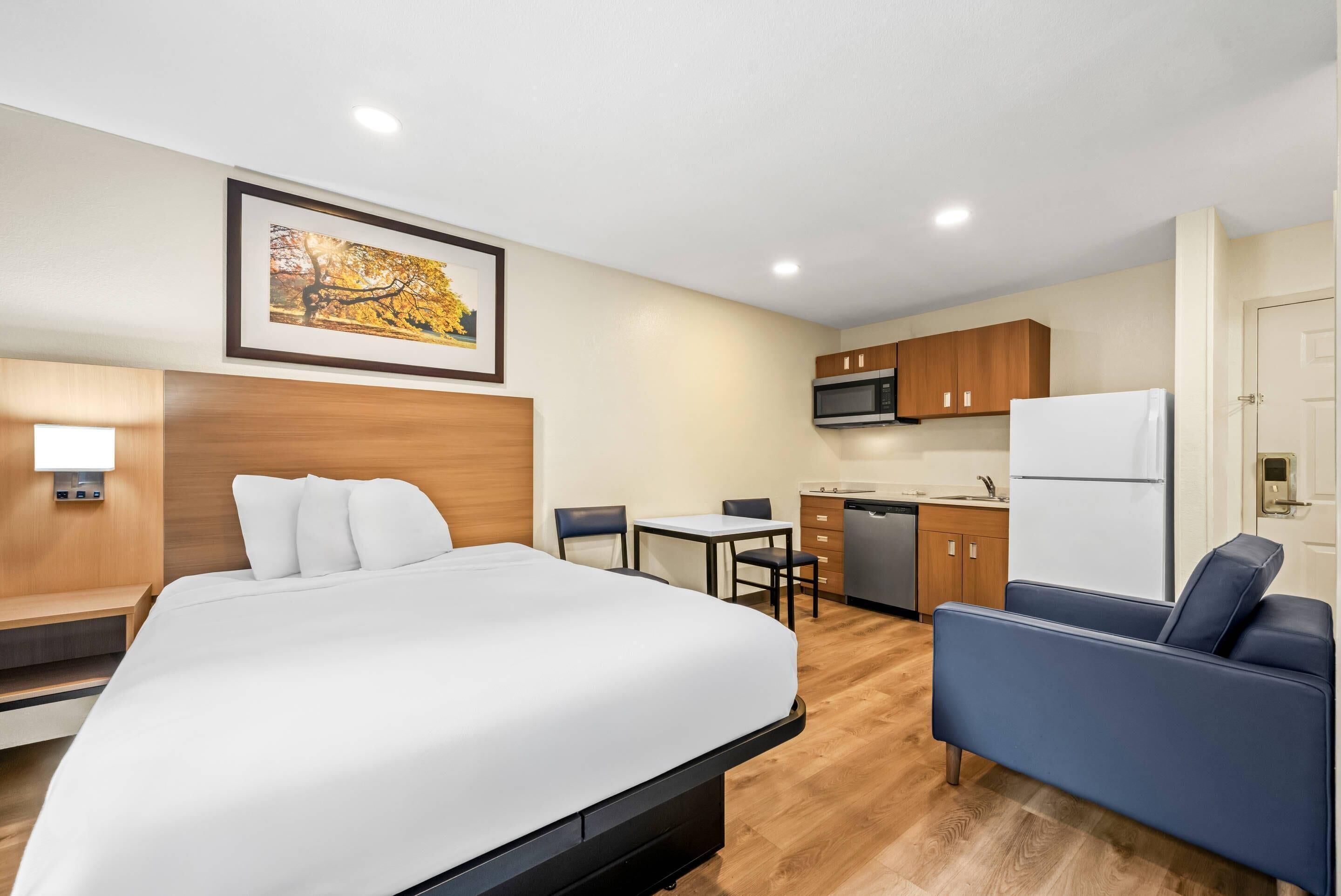 A hotel room with a bed, a desk, a chair, a sofa, a kitchen area, and a painting on the wall.