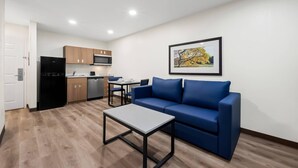 A modern living room with a blue sofa, a coffee table, and a kitchen area with wooden cabinets and appliances. - MainStay Suites Raleigh North (Raleigh)