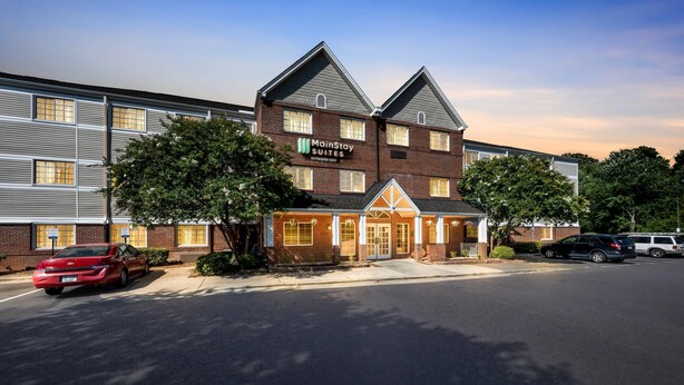 A MainStay Suites hotel with a brick facade, large windows, and a parking lot with cars. - MainStay Suites Raleigh North (Raleigh)