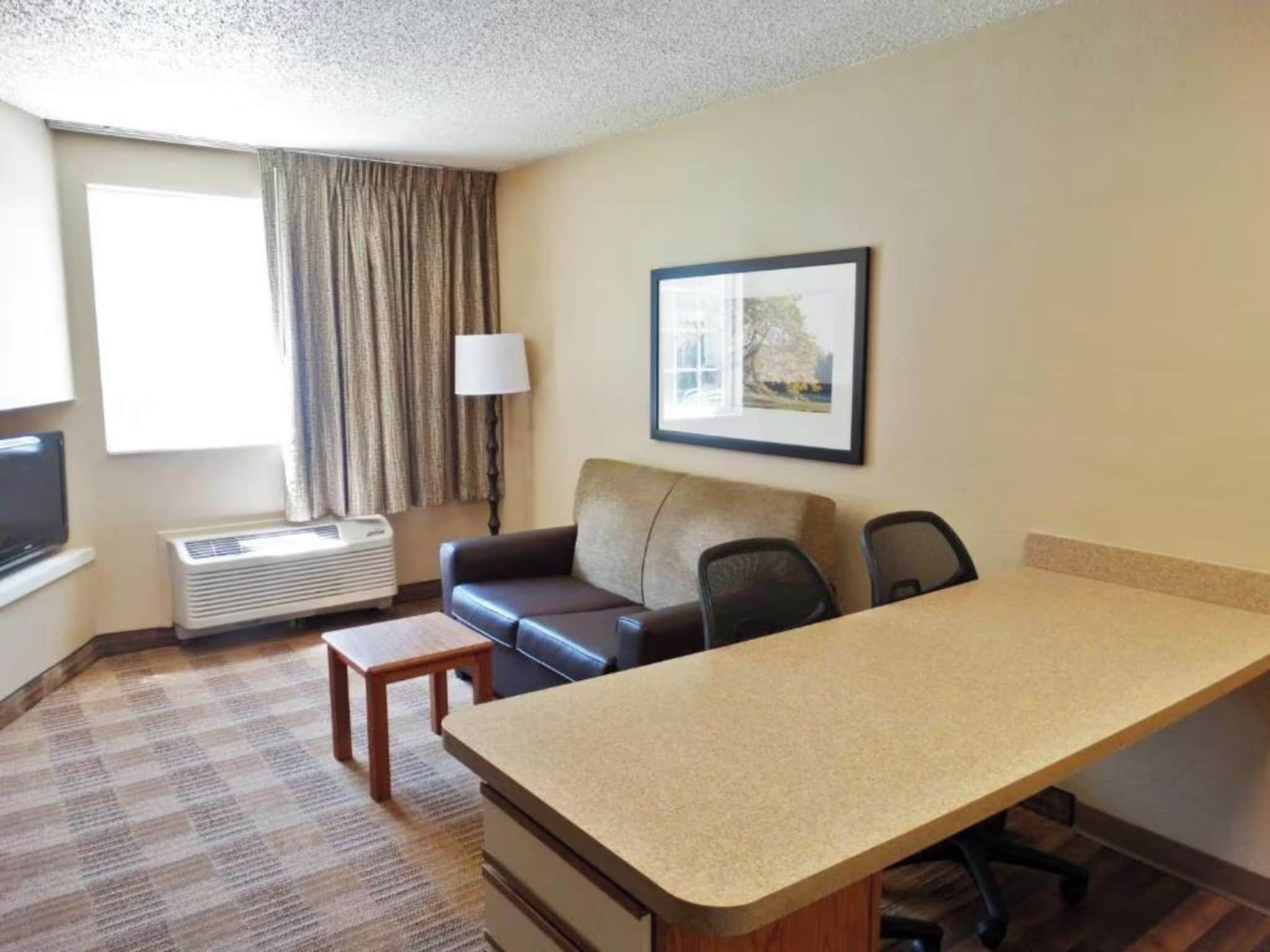 A hotel room with a sofa, a small table, a desk, a chair, a lamp, a framed picture, and a window with curtains.