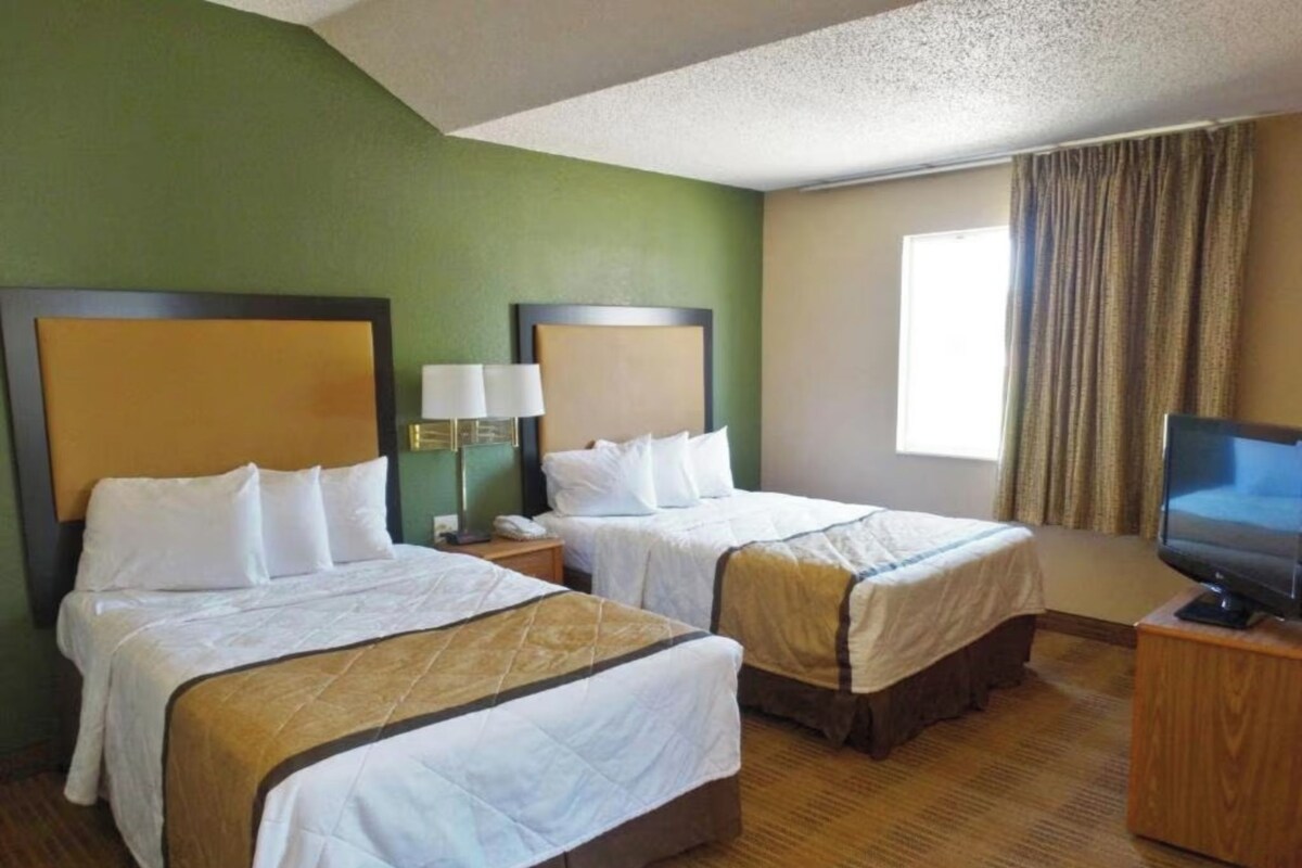 A hotel room with two beds, a television on a wooden stand, and a window with curtains.