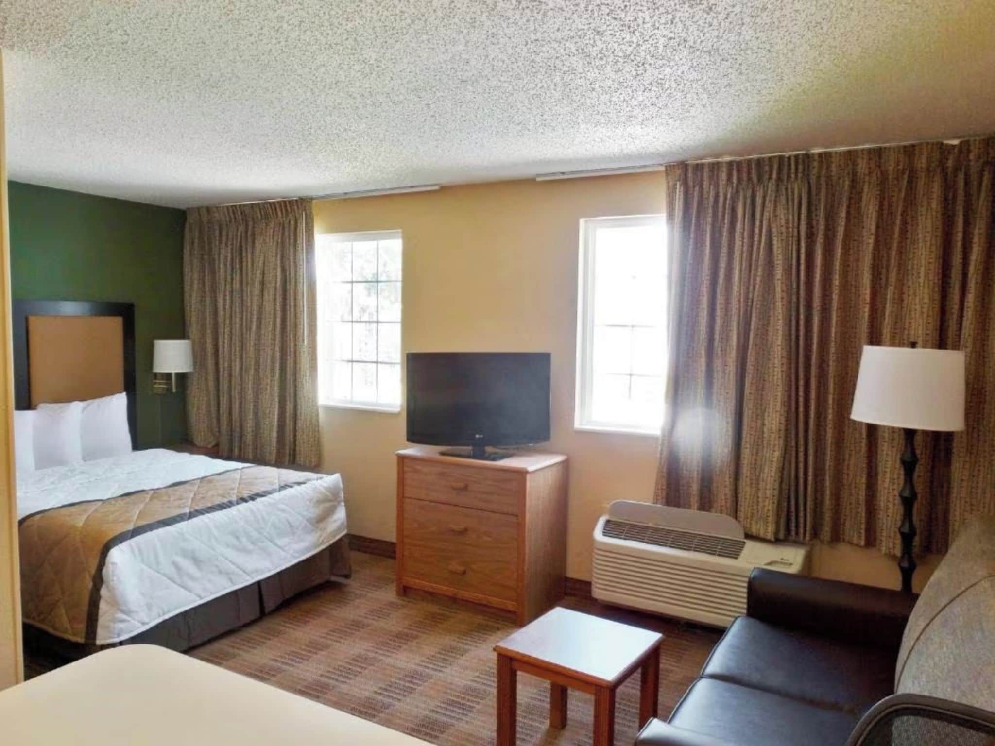 A hotel room with a bed, a TV on a wooden dresser, a sofa, and an air conditioning unit.