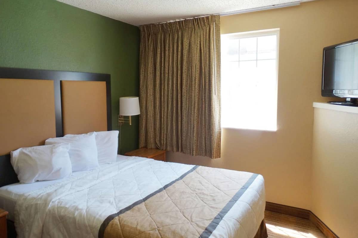 Una habitación de hotel con cama, televisor, ventana con cortinas y mesita de noche con lámpara.