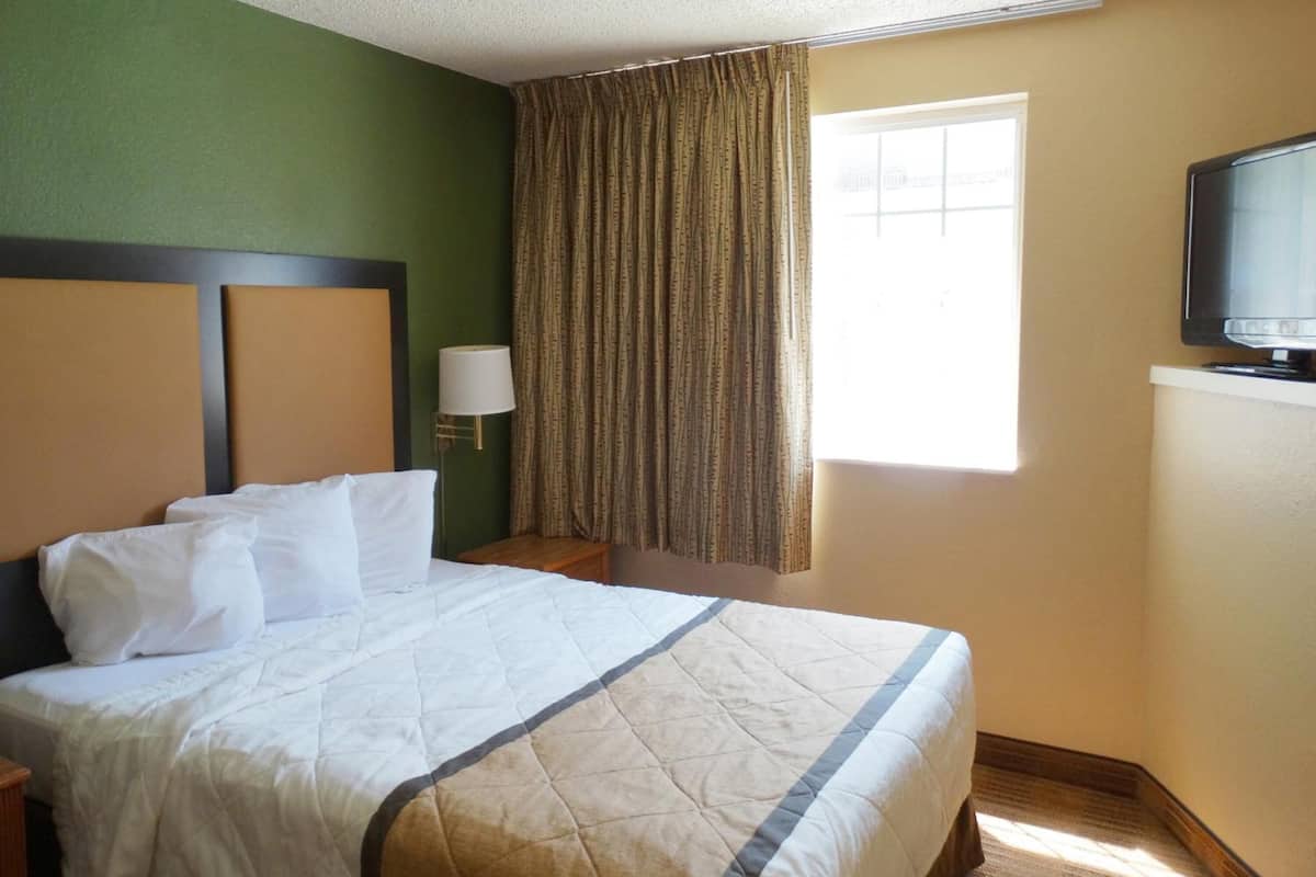 Una habitación de hotel con una cama, televisión, ventana con cortinas y una lámpara.