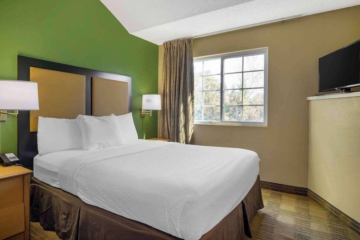 Quarto de hotel com uma cama grande, duas luminárias de cabeceira, televisão e uma janela com cortinas.