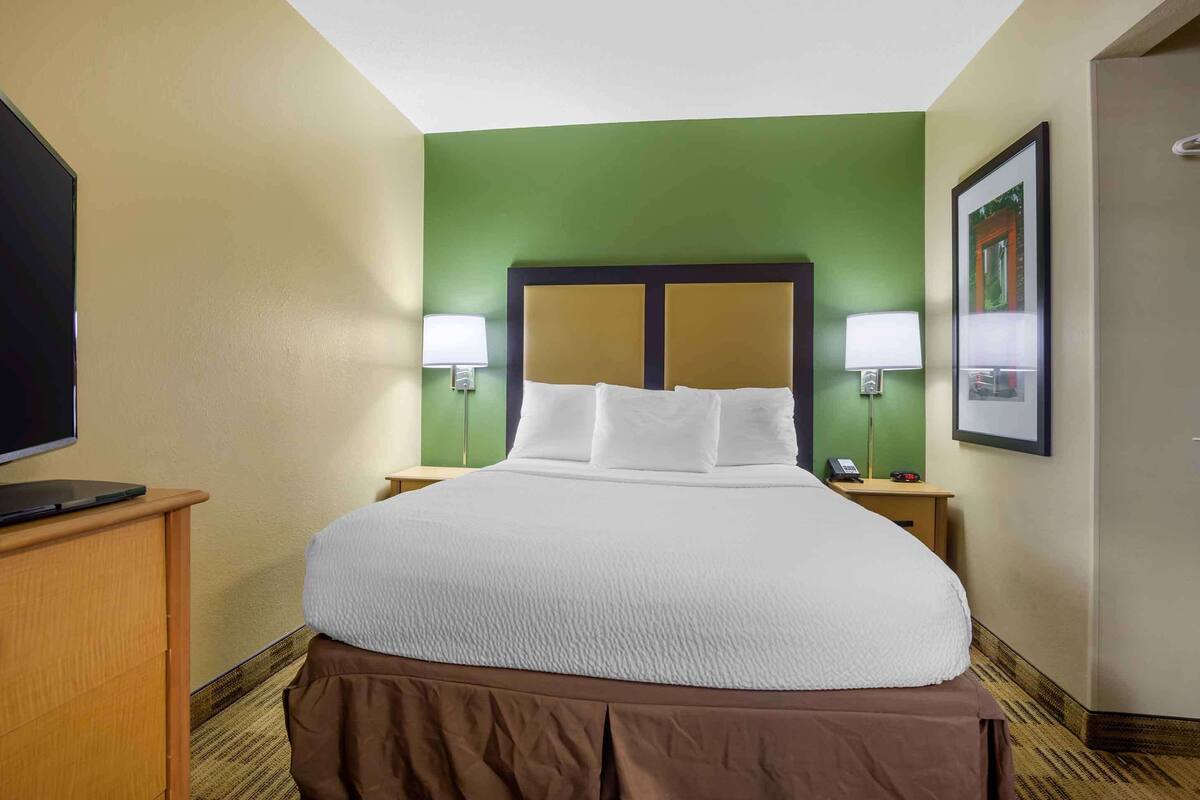 A hotel room with a large bed, two bedside lamps, a flat-screen TV, and a wooden nightstand.